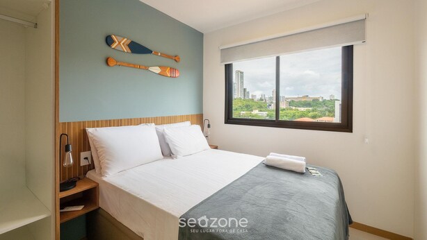Quarto de hotel com cama, mesa de cabeceira, luminária, janela com vista para a cidade e dois pranchas de remo na parede.