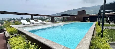 Een dakzwembad met helderblauw water, omringd door groen en met uitzicht op een stadsgezicht met bergen in de verte.