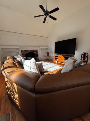 Una sala de estar con un sofá de cuero marrón, un televisor de pantalla plana, chimenea y ventilador de techo.