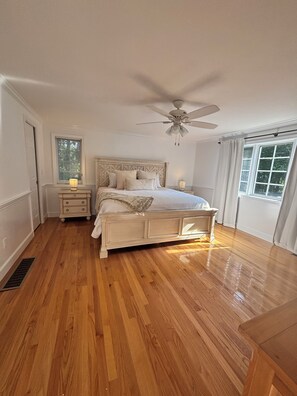 Un dormitorio con piso de madera, una cama con cabecera, una mesita de noche, una ventana con persianas y un ventilador de techo.
