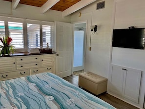 Ein Schlafzimmer mit Bett, Kommode, Sitzbank, Fernseher und einem Fenster mit Jalousien.