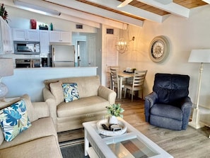 Ein Wohnzimmer mit Sofa, Sessel und Couchtisch. Es gibt einen Essbereich mit Stühlen und Tisch. Im Hintergrund ist eine Küche erkennbar.