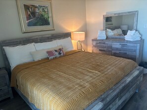 Un lit avec une tête de lit en bois, une lampe de chevet, une commode avec des serviettes pliées et un tableau encadré au mur.