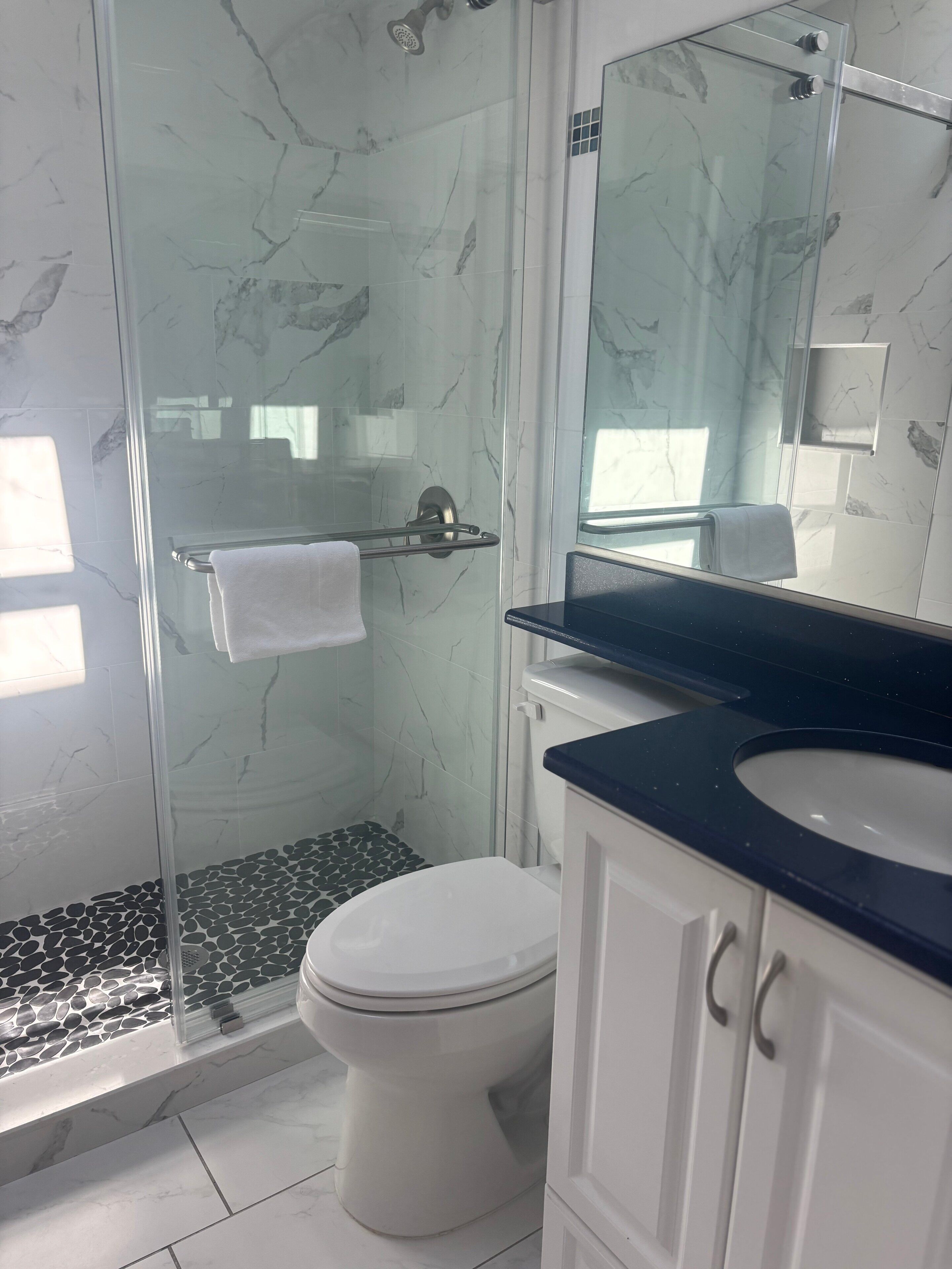Une salle de bain avec une douche à parois de verre, des toilettes blanches, un lavabo sur un plan de travail noir et un miroir au-dessus du lavabo.