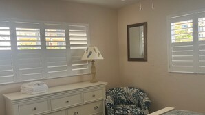 A bedroom with a bed, a dresser with drawers, a chair, a lamp, and a window with blinds. - Gulf-front 1BR Siesta Key condo with king bed and sleeper sofa. (Siesta Key)