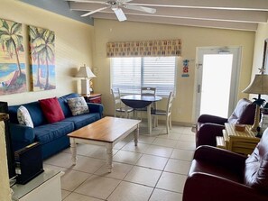 A living room with a blue sofa, a wooden coffee table, a red armchair, a dining table with chairs, and a ceiling fan. - Gulf-front 1BR Siesta Key escape with king bed and sleeper sofa. (Siesta Key)