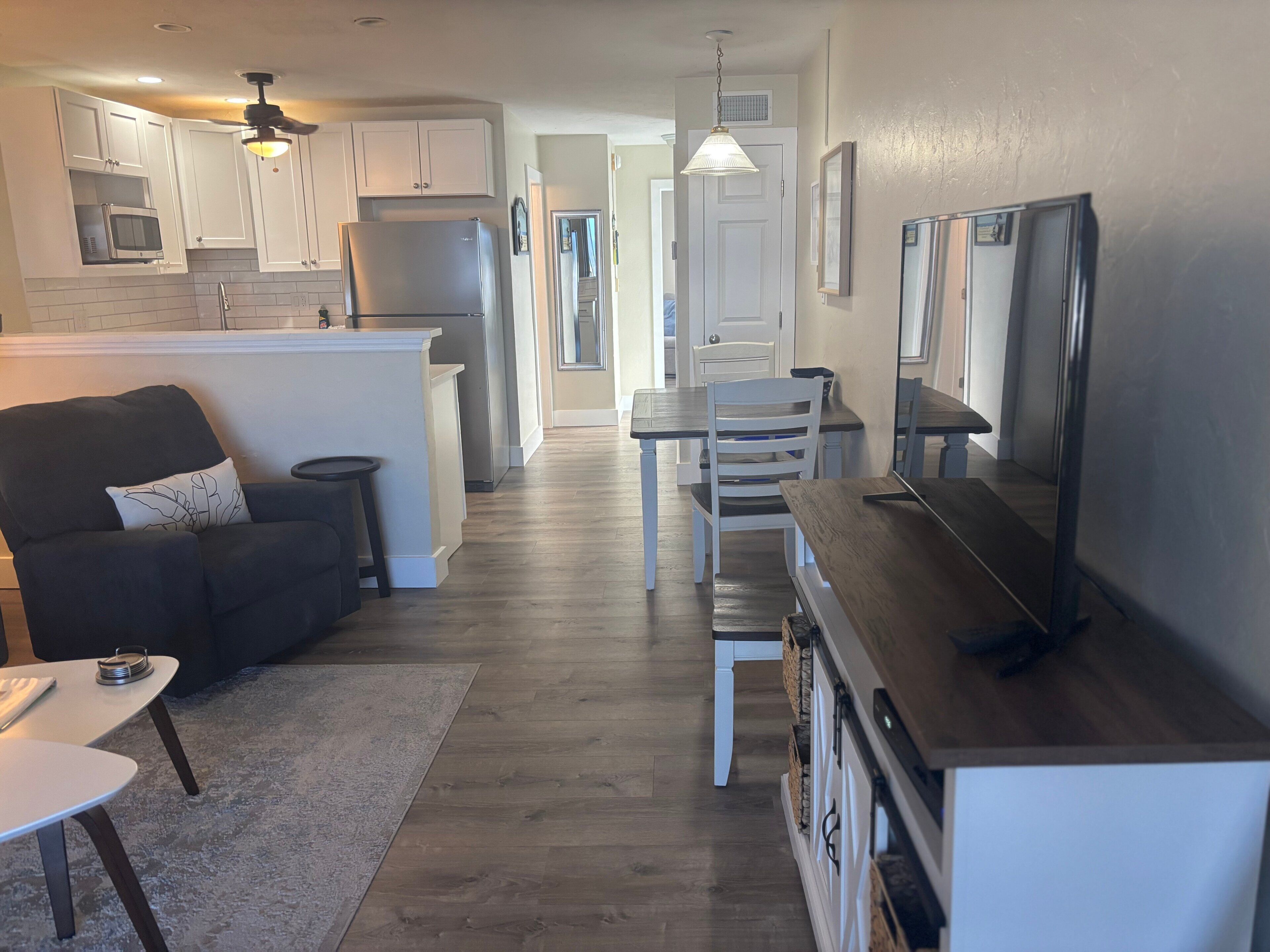 A modern kitchen with white cabinets, a microwave, and a refrigerator. There is a dining area with a table and chairs, and a living room with a sofa and a TV.