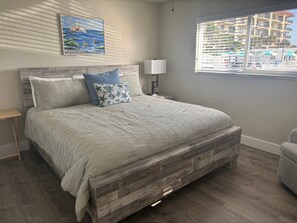 A bedroom with a wooden bed frame, a nightstand, a lamp, and a window with a view of buildings. - Gulf-front 1BR Siesta Key condo with king bed and sleeper sofa. (Siesta Key)
