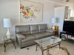 A living room with a grey sofa, two glass tables, a flat-screen TV, and a bookshelf. - SR-10 Gulf Front 1bed/1bath (Siesta Key)
