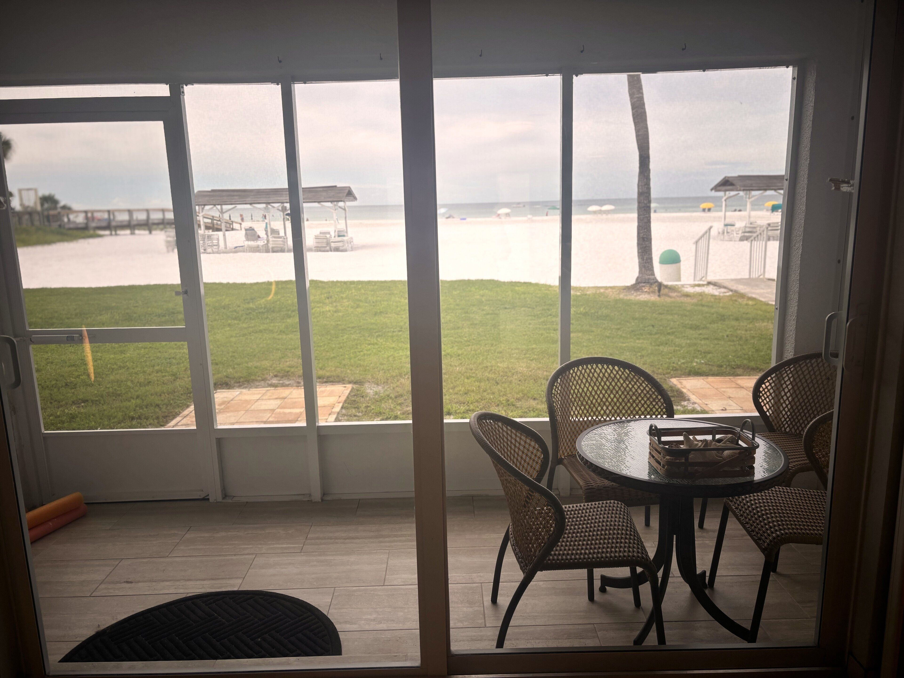 Una habitación con puerta de vidrio que da a un patio con mesa y sillas, con vista a una playa con sombrillas y un muelle.