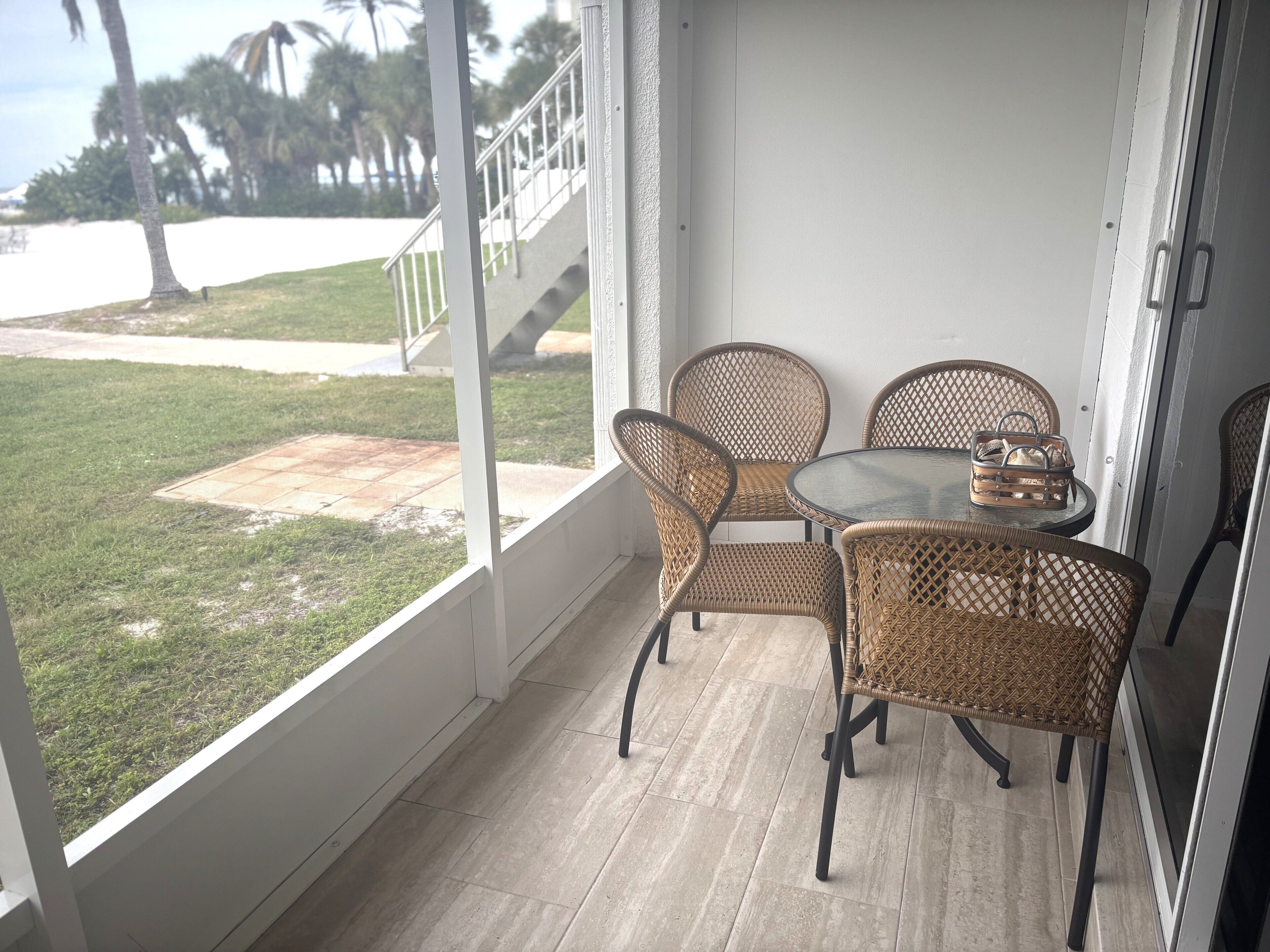 Un balcón con mobiliario de mimbre y una mesa de vidrio, con vista a una zona verde y palmeras.