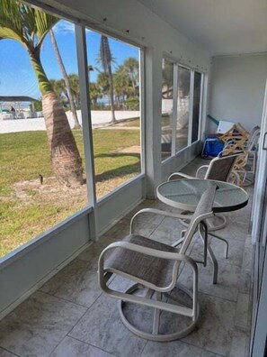 A covered outdoor seating area with a glass table and chairs, a view of palm trees, and a clear sky. - SR-12A Gulf Front 1/1 (Siesta Key)