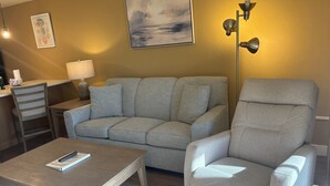 En stue med sofa, lænestol, sofabord, lampe og et billede på væggen.