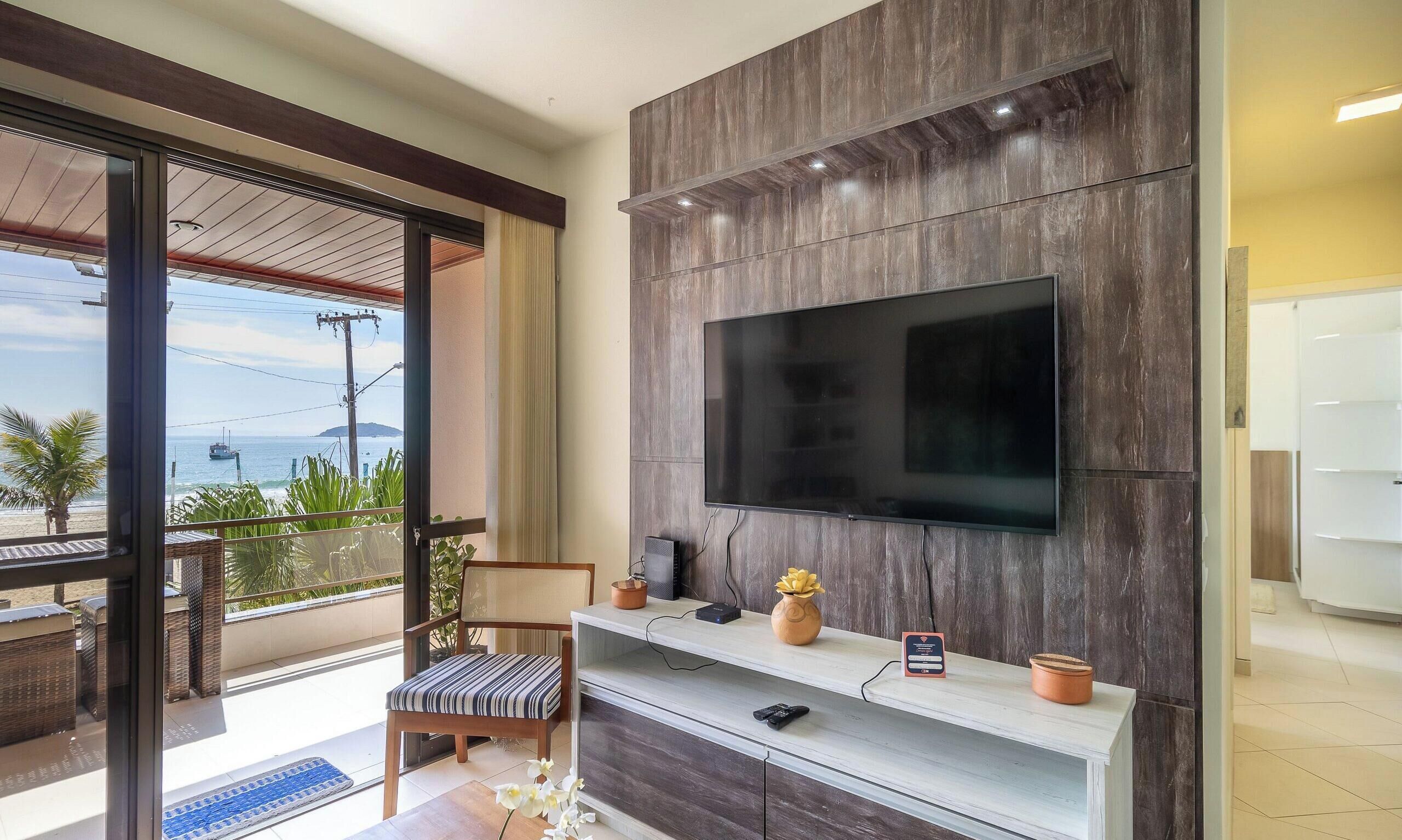 Una sala moderna con un televisor de pantalla plana grande montado en una pared de madera, una consola blanca con objetos decorativos y vistas al océano a través de una puerta corrediza de vidrio.