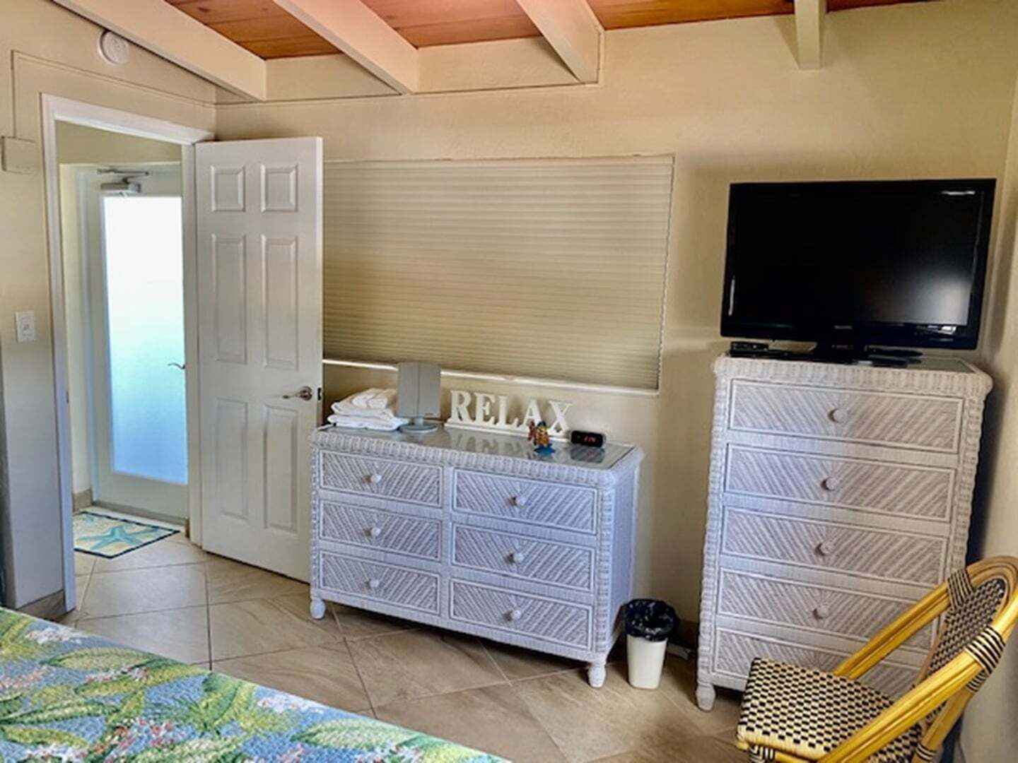 A room with a bed, a white dresser with drawers, a television on a stand, a wicker chair, and a door leading to another room.