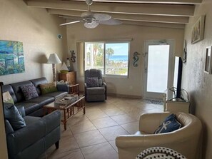 Ein Wohnzimmer mit einer Couch, einem Sessel und einem Couchtisch. Es gibt einen Deckenventilator, ein Fenster mit Aussicht und eine Tür, die nach draußen führt.