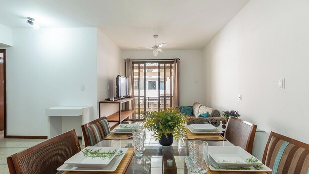 A dining area with a table set for four, chairs, a television on a stand, and a view through a large window. - Beautiful Apartment Right on the Sand Ilh0107 (Florianópolis)