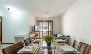 A dining area with a table set for four, chairs, a television on a stand, and a view through a large window.