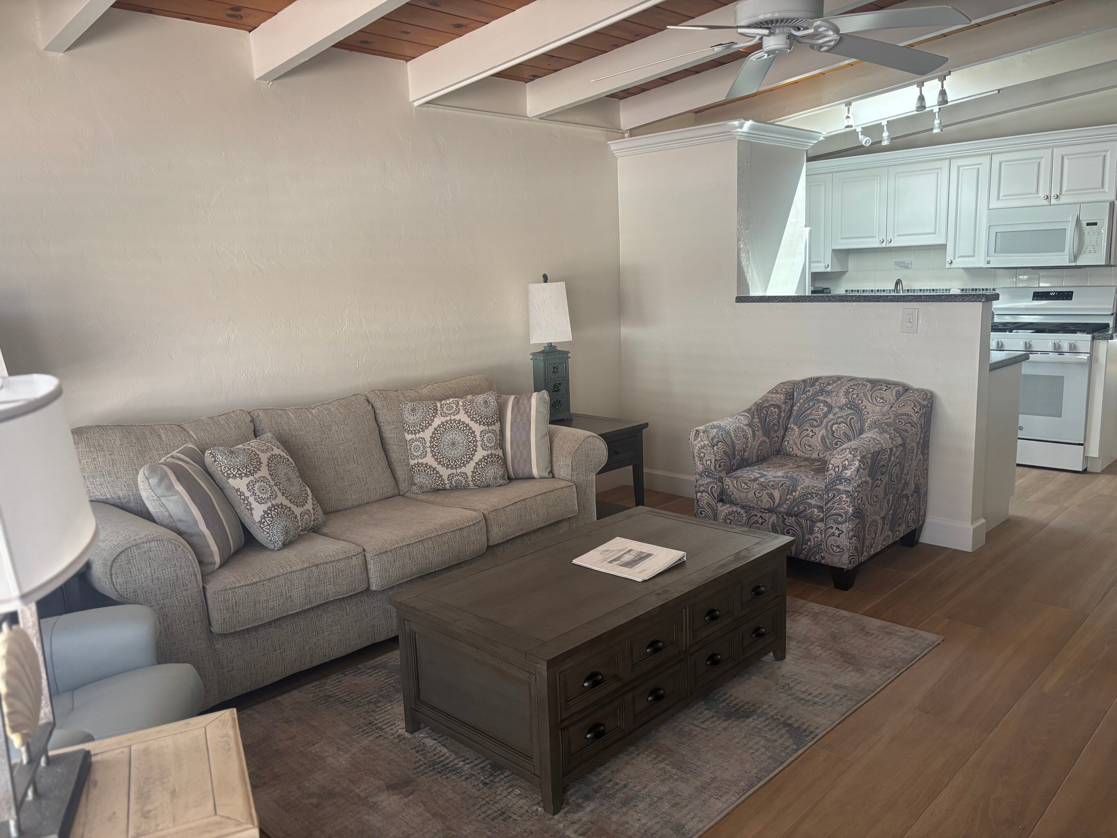 Ein Wohnzimmer mit Sofa, Sessel, Couchtisch und einer Küche im Hintergrund.