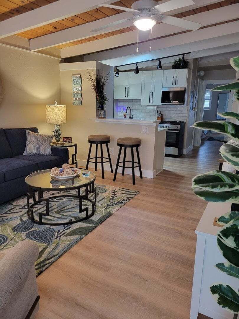 Una sala moderna con sofá, mesa de centro y taburetes, que da a una cocina con armarios blancos y una isla central.