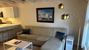 A living room with a sofa, a coffee table, a floor lamp, and a painting on the wall. - SR-23 Lovely 1/1 Unit (Siesta Key)
