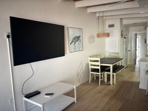 A modern living space with a flat-screen TV mounted on the wall, a dining area with a wooden table and chairs, and a kitchen area with white cabinets and a hanging light fixture. - SR-23 Lovely 1/1 Unit (Siesta Key)