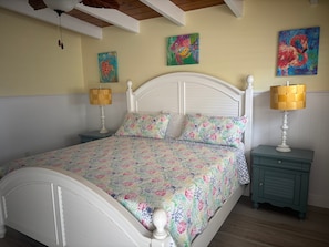 A bedroom with a white bed, floral bedding, two bedside lamps, and a teal nightstand. - SR- North Court Unit (Siesta Key)