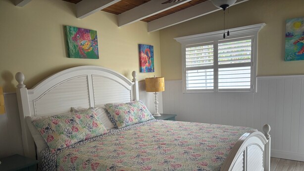 A bedroom with a white bed, floral bedding, a nightstand, a lamp, and artwork on the walls. - SR- North Court Unit (Siesta Key)