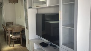Una sala de estar moderna con un televisor de pantalla plana grande montado en un mueble blanco con puertas de cristal, una mesa de comedor con sillas y una lámpara colgante.