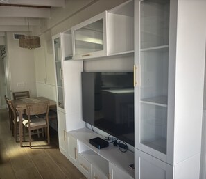 Ein modernes Wohnzimmer mit großem Flachbildfernseher, der an einem weißen Schrank mit Glastüren befestigt ist, einem Esstisch mit Stühlen und einer Pendelleuchte.