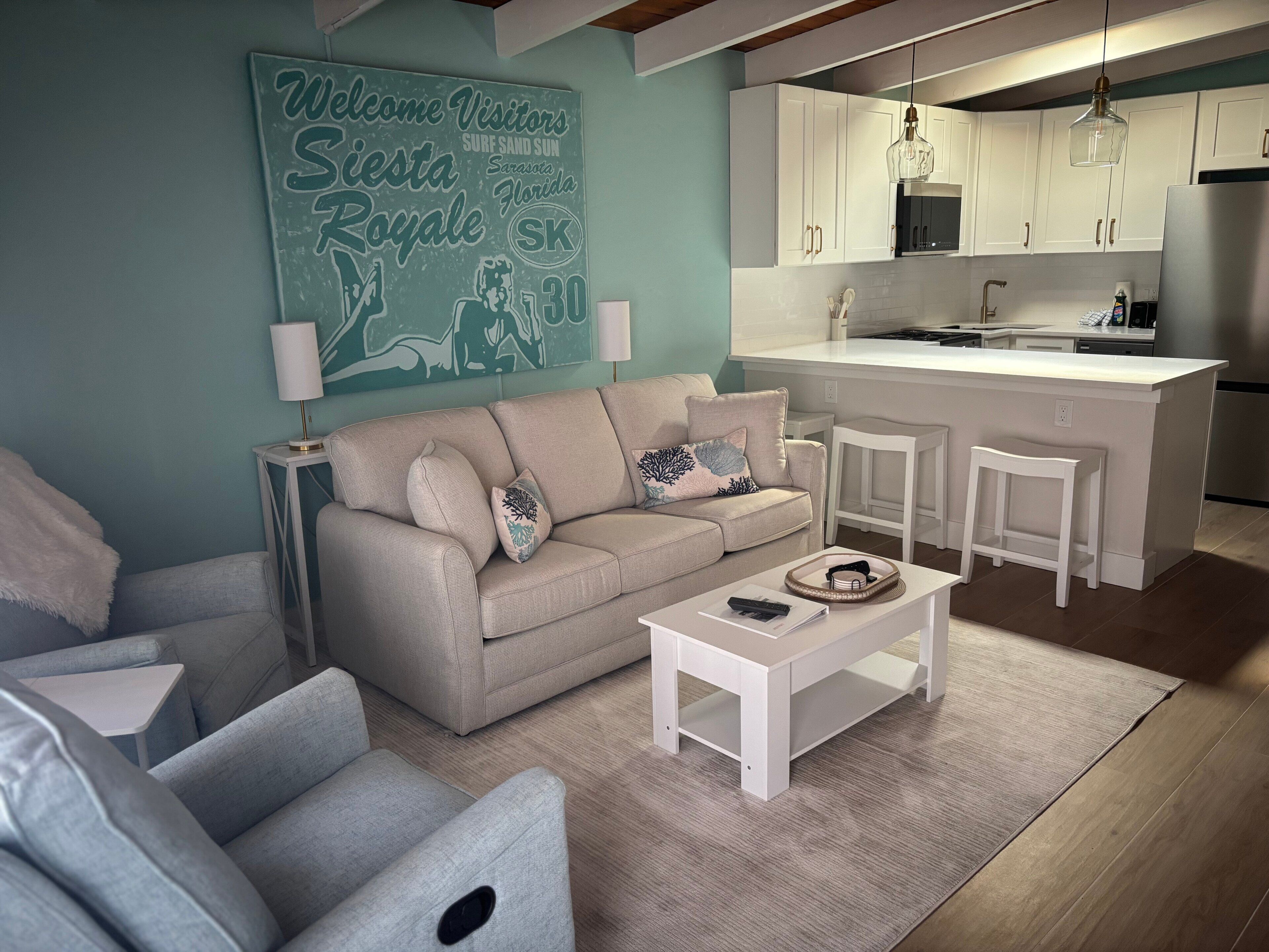 A modern living room with a sofa, armchairs, and a coffee table. A kitchen with white cabinets and a stainless steel refrigerator is visible in the background.