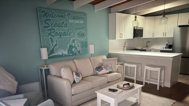 Una sala de estar moderna con un sofá, sillones y una mesa de centro. Se ve una cocina al fondo con armarios blancos y un refrigerador de acero inoxidable.