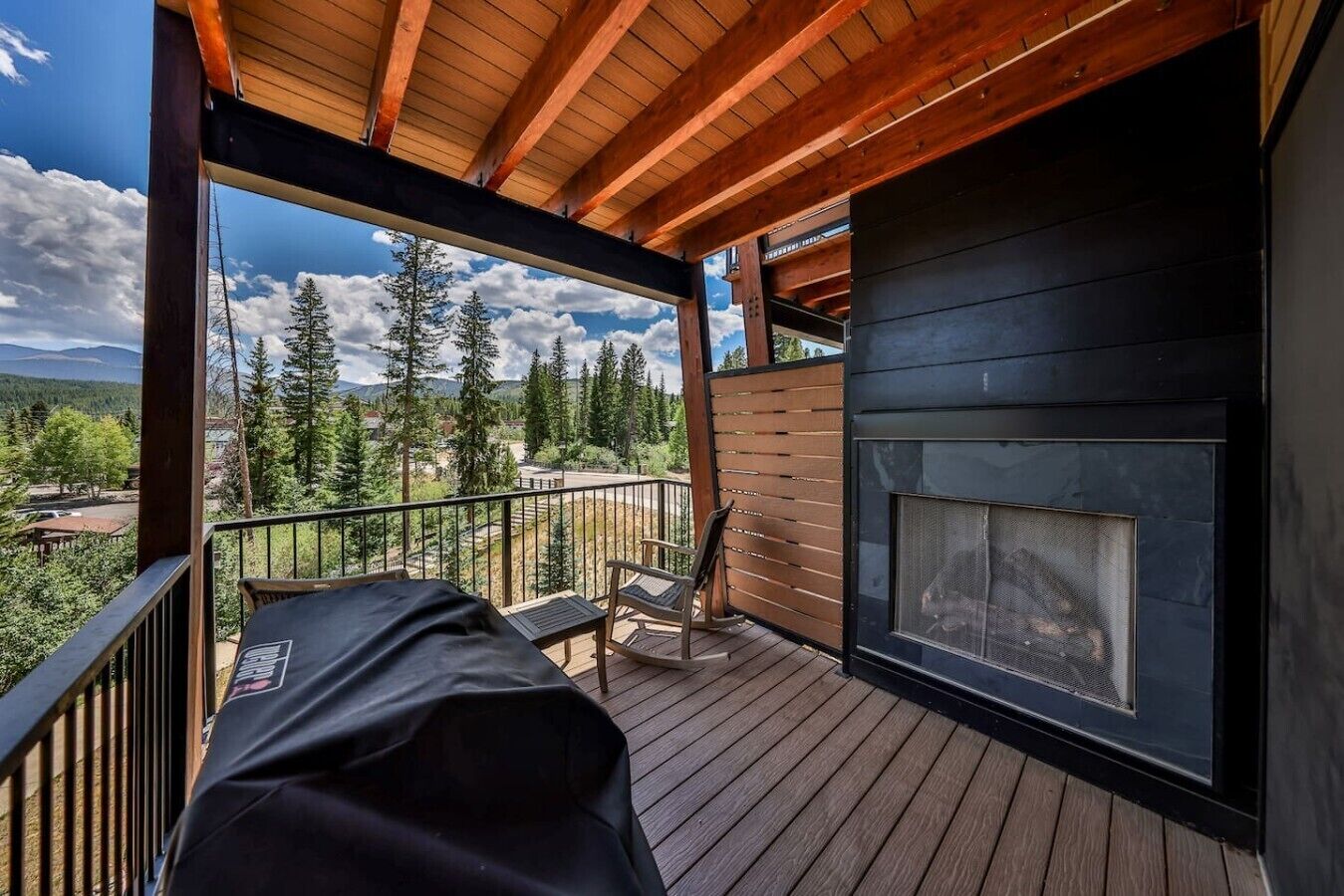 A wooden deck with a fireplace, outdoor seating, and a view of a forest and mountains.