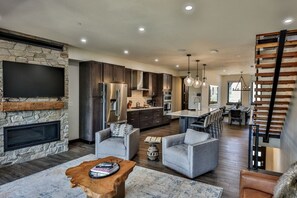 A spacious living area with a stone fireplace, modern kitchen, and a dining area with a staircase. - Hot Tub | Mountain Views | Walk To Downtown (Winter Park)