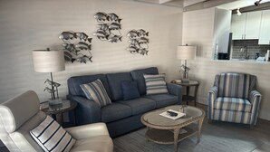 Ein modernes Wohnzimmer mit Sofa, Sessel und Couchtisch. Im Hintergrund ist eine Küchenzeile erkennbar.