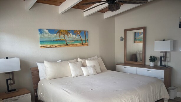 Une chambre avec un grand lit, un ventilateur de plafond, un tableau représentant une plage, un miroir et une table de chevet avec une lampe.