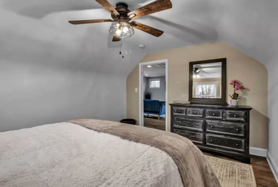 Une chambre à coucher avec un ventilateur de plafond, un lit recouvert d’une couverture blanche, une commode et un miroir.