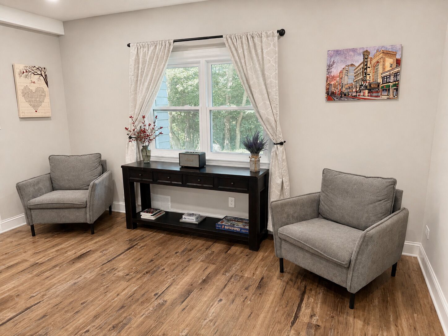 A living room with two grey armchairs, a dark wooden console table with books, a window with white curtains, and a cityscape painting on the wall.