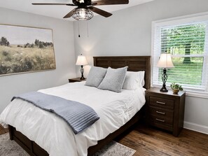A bedroom with a bed, two bedside tables with lamps, a ceiling fan, a window with blinds, and a painting on the wall.