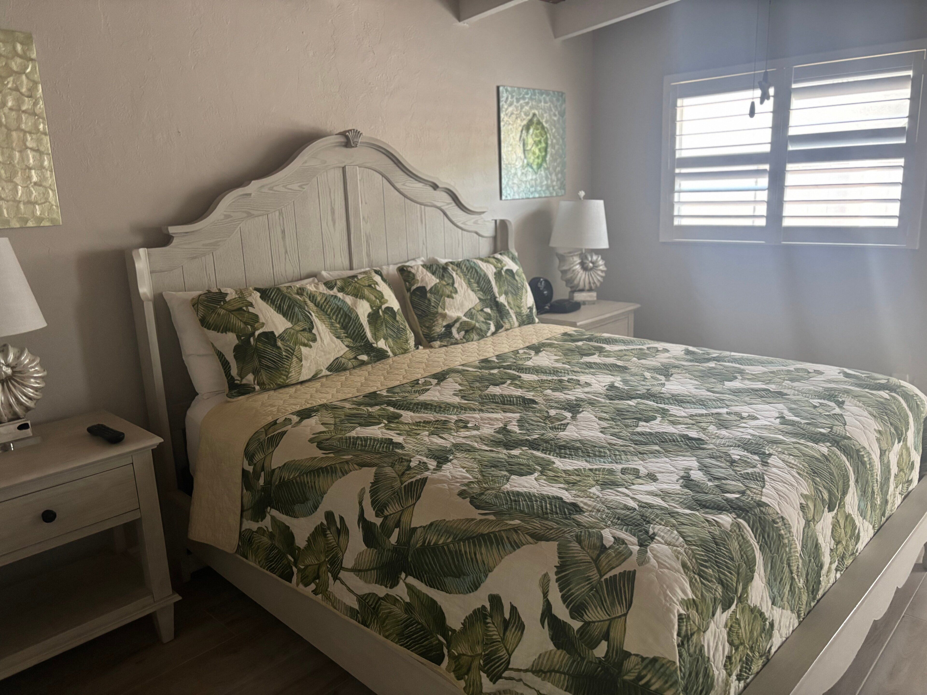 Ein Schlafzimmer mit einem Bett, Nachttischen, einer Lampe, einem Wandbild und einem Fenster mit Jalousien.
