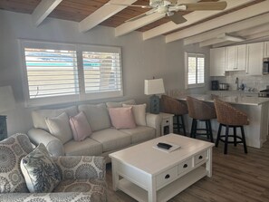 A living room with a sofa, patterned armchair, and a coffee table. There is a kitchen area with white cabinets and a bar stool. - SR-40, Lovely Courtside Unit (Siesta Key)