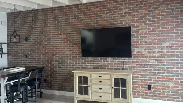 A room with a brick wall, a flat-screen TV mounted above a wooden cabinet, and a dining area with chairs and a table. - SR-45 - 1BR Siesta Key condo with king bed, sleep sofa, and courtyard views. (Siesta Key)