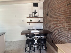 A dining area with a wooden table and four black chairs, a hanging light fixture, and a brick wall. - SR-45 - 1BR Siesta Key condo with king bed, sleep sofa, and courtyard views. (Siesta Key)