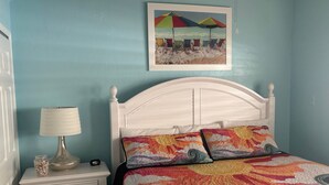 A bedroom with a white bed, colorful pillows, a framed beach painting, a bedside lamp, and a nightstand with a jar and a clock. - SR-46 -Cozy 1BR Siesta Key apartment with king bed and courtyard views. (Siesta Key)