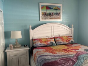 A bedroom with a white bed, colorful pillows, a framed beach painting, a bedside lamp, and a nightstand with a jar and a clock. - SR-46 -Cozy 1BR Siesta Key apartment with king bed and courtyard views. (Siesta Key)