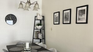 Un área de comedor con una mesa de madera oscura, individuales grises, sillas blancas, un reloj de pared y tres cuadros enmarcados.