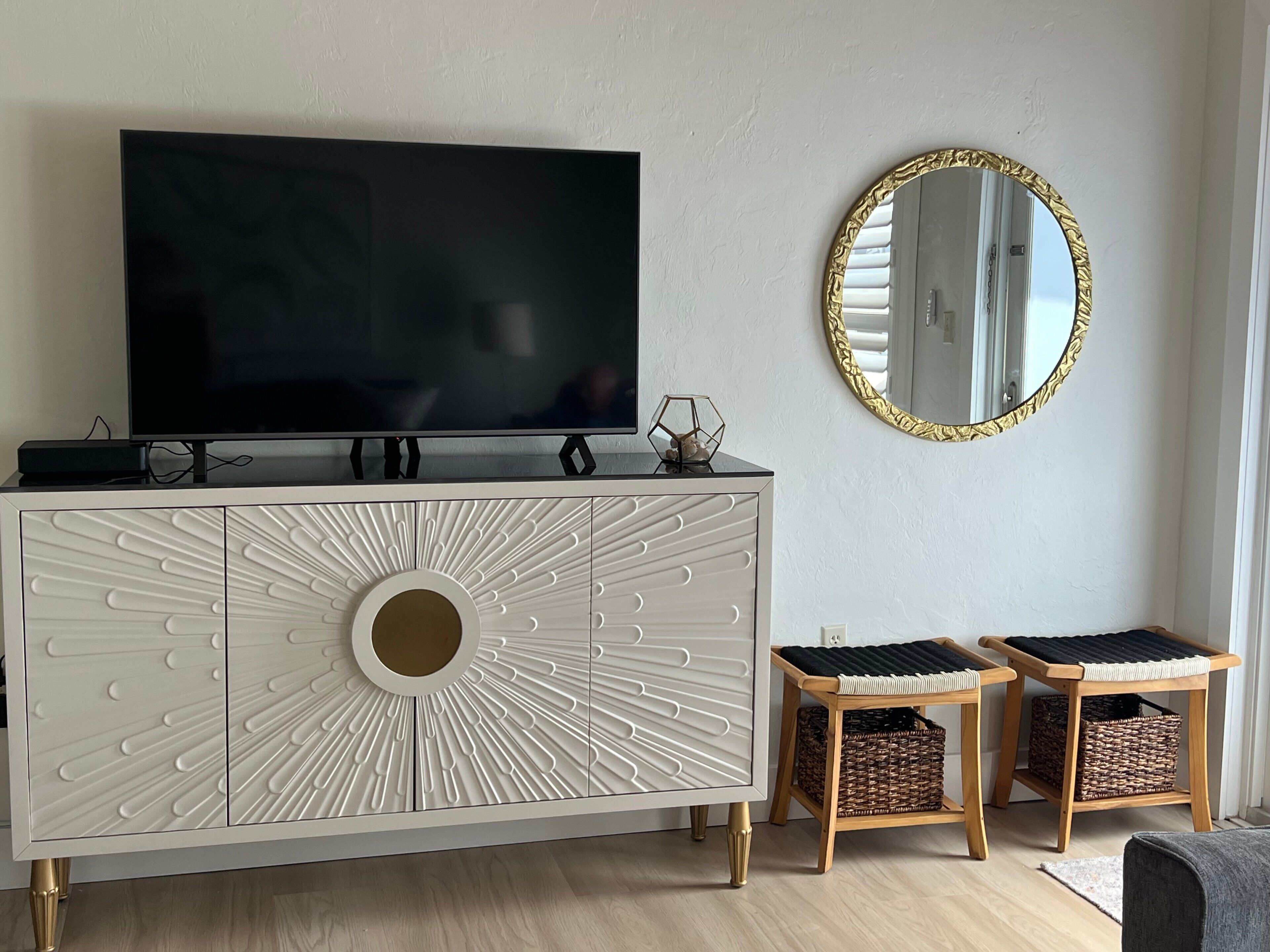 Uma sala de estar moderna com uma televisão de tela plana em um armário branco, um espelho redondo com moldura dourada e duas cestas de vime sobre banquinhos de madeira.