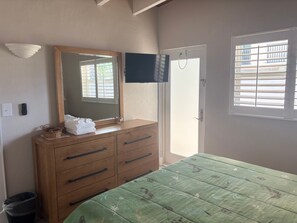 A bedroom with a wooden dresser, a bed with a floral bedspread, a mirror, a television on the wall, and a window with blinds. - SR59 -1BR Siesta Key condo with courtyard view, queen bed, and sleeper sofa. (Siesta Key)