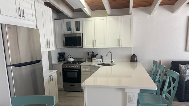 A modern kitchen with white cabinetry, stainless steel appliances, and a central island with teal chairs. - SR-50 -1BR 1BA Courtyard Unit (Siesta Key)