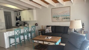 A modern kitchen with white cabinets, a dining area with teal chairs, a wooden coffee table, and a gray sofa. - SR-50 -1BR 1BA Courtyard Unit (Siesta Key)